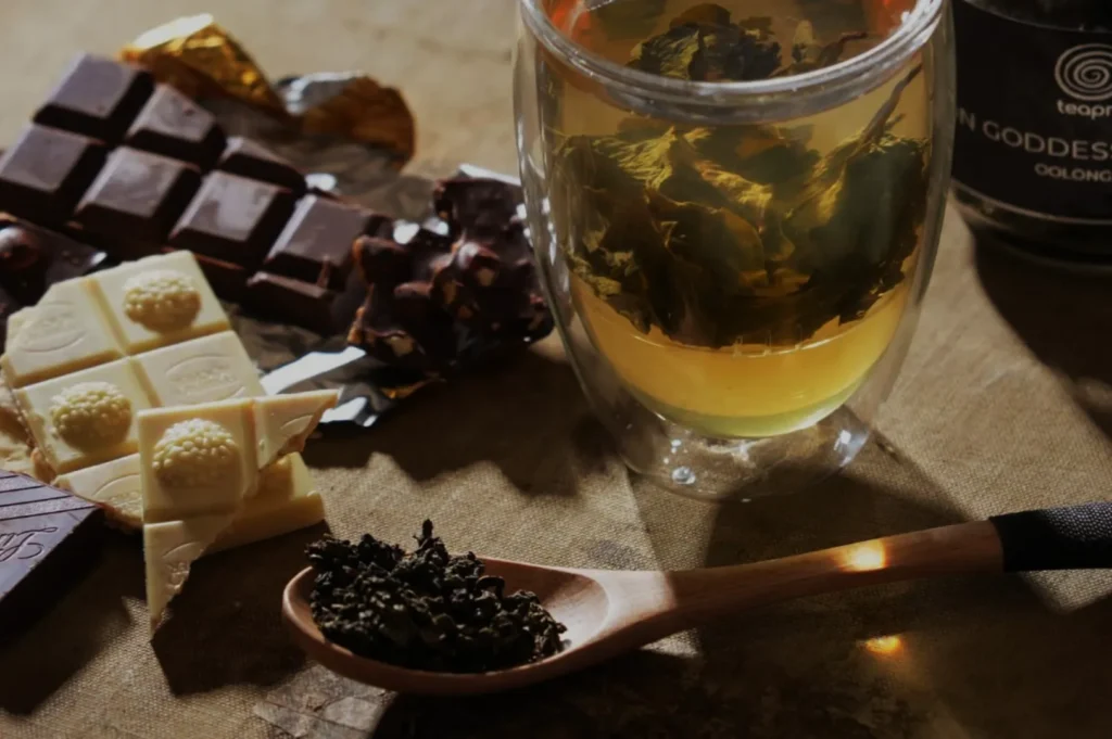A glass of tea with floating leaves is surrounded by an assortment of chocolate pieces and a wooden spoon holding loose tea leaves on a textured surface.