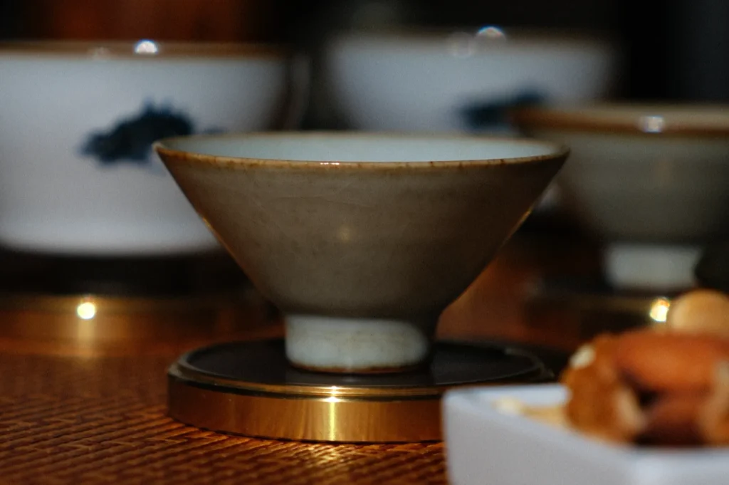 A ceramic teacup on a brass coaster sits on a textured wooden table, with blurred background cups. Warm, intimate atmosphere.
