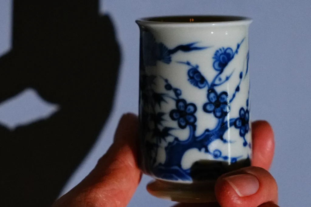A hand holding a small porcelain cup decorated with blue floral patterns, casting a shadow on the wall.