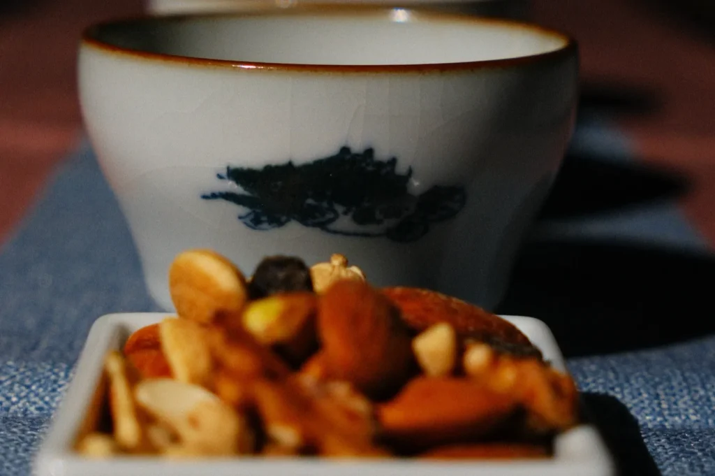 A small white cup with a blue pattern is in the background. In the foreground, a square dish holds mixed nuts. The scene is dimly lit, creating a cozy mood.