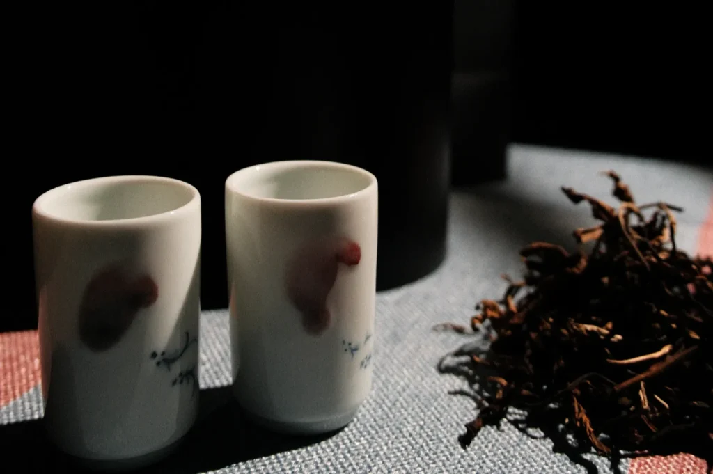 Two small, decorative ceramic cups next to a pile of dried tea leaves, set against a dark background.