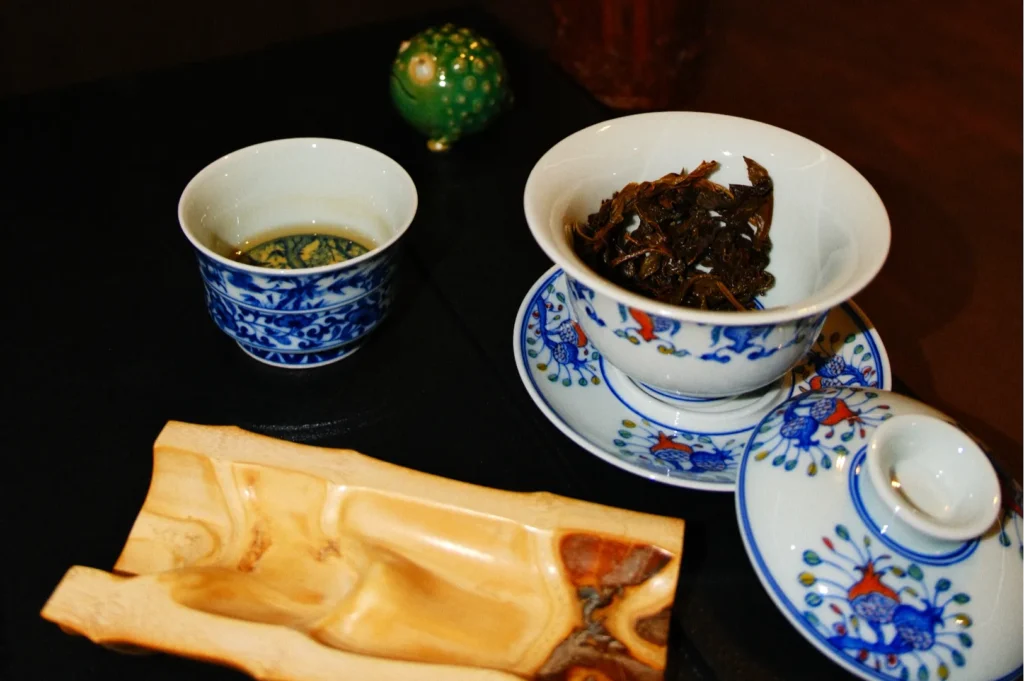 A traditional tea setup featuring a blue and white patterned teacup, a bowl of dried tea leaves, and a wooden tray.