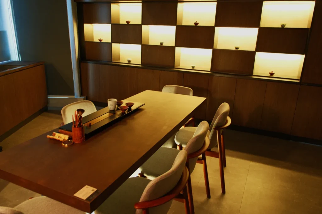 A modern dining area with a wooden table and light-colored chairs, featuring a softly lit shelf with decorative items in the background.