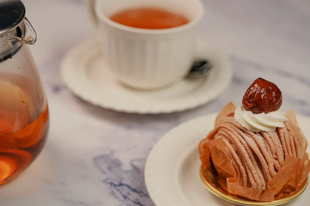 A delicate Mont Blanc dessert topped with chestnut and cream sits on a white plate. Beside it, a cup and a pot of tea on a marble surface create an elegant, cozy vibe.