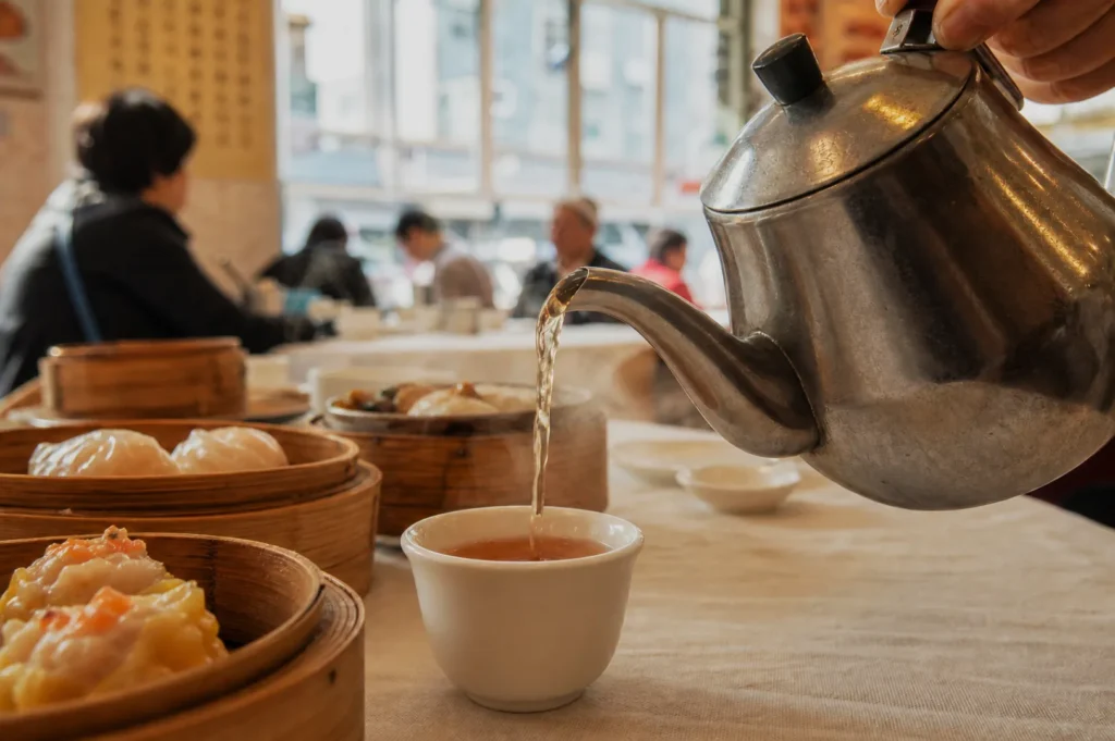 A teapot pours tea into a cup on a table set with dim sum baskets. Blurred diners and large windows in the background create a warm, lively atmosphere.