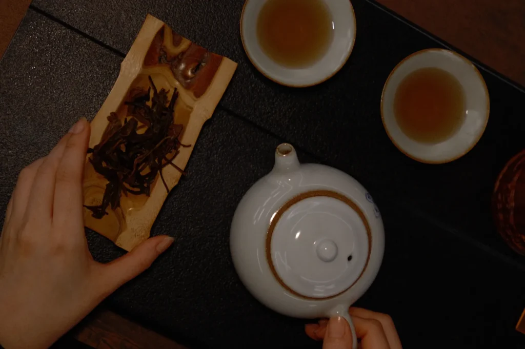 A person's hand pours tea from a white teapot onto two cups, with dried tea leaves on a piece of paper nearby.