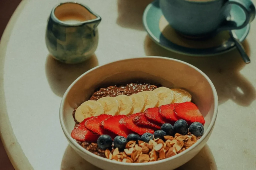 A bowl of chocolate oatmeal topped with banana slices, strawberries, blueberries, and granola on a table. A latte and milk jug are in the background.