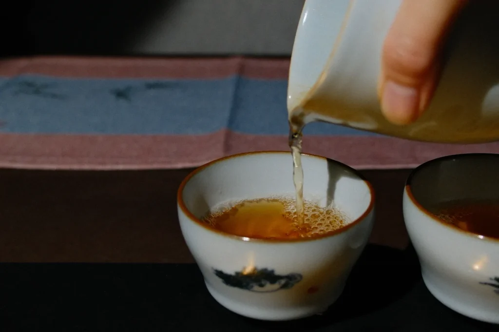 A hand is pouring tea from a ceramic teapot into a small white cup. The cup has a decorative design, and there is another empty cup beside it. A colorful cloth is visible in the background, featuring light blue and pink stripes.
