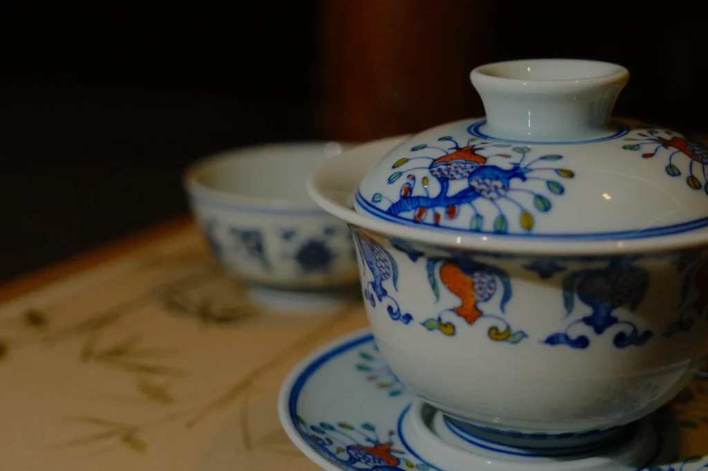 Porcelain teacup with lid and saucer featuring blue and orange floral patterns in focus, with a softly blurred background cup on a bamboo mat.