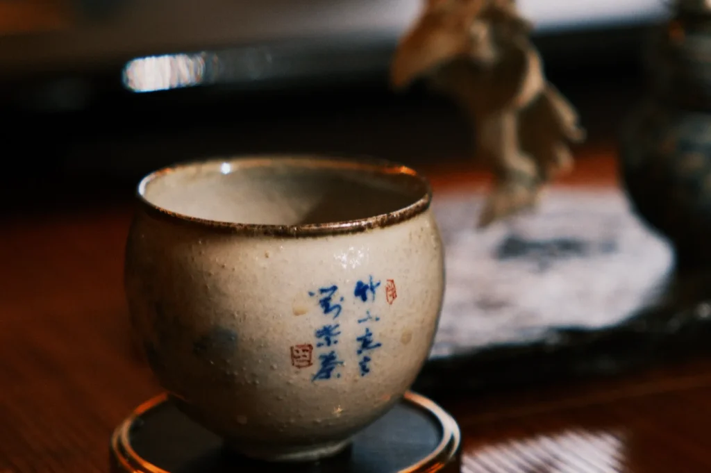 Ceramic cup with blue Chinese characters and a rustic glaze sits on a wooden table. Background is blurred, creating a calming, contemplative mood.
