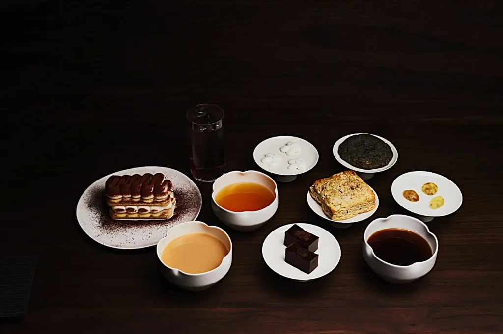 A visually appealing arrangement of desserts and drinks on a dark table. Desserts include tiramisu, chocolate cubes, and pastries, paired with tea and coffee.