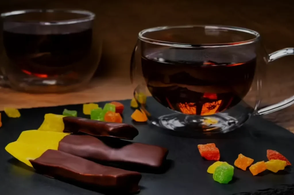 A glass cup of dark tea sits on a slate surface, surrounded by colorful candied fruit and chocolate-covered slices. Another cup is blurred in the background.