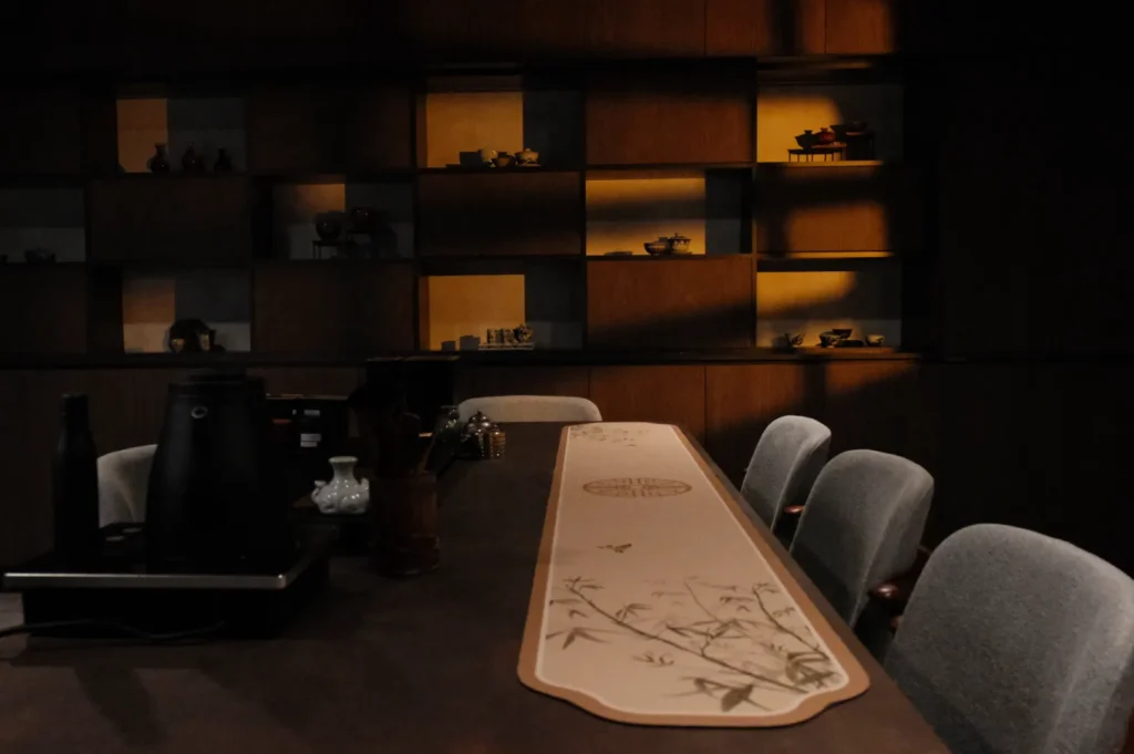 A dimly lit room with a long table featuring a decorative runner, surrounded by several chairs. Behind the table, there are shelves with various objects displayed.