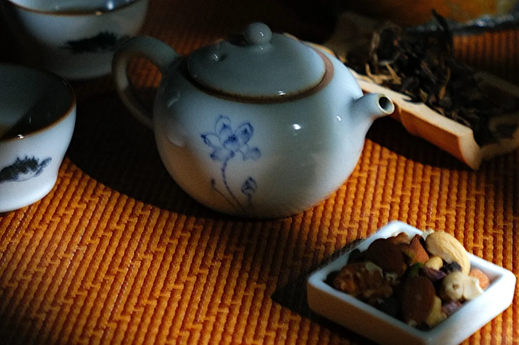 A small, white ceramic teapot with blue floral design sits on a woven mat. Nearby are teacups and a tray with assorted nuts and dried tea leaves, creating a cozy, serene atmosphere.