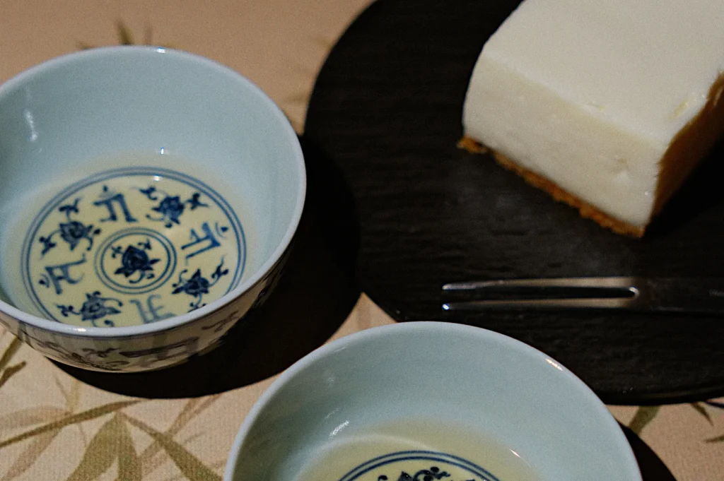 Two decorative bowls with intricate blue patterns sit beside a slice of creamy cake on a dark plate. A fork is placed near the cake, creating a serene and inviting setting.