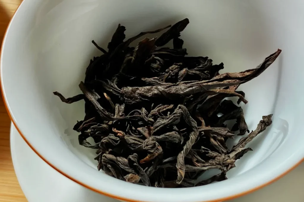 Dried black tea leaves arranged in a white porcelain bowl with a subtle gold rim, placed on a wooden table, conveying a sense of warmth and simplicity.