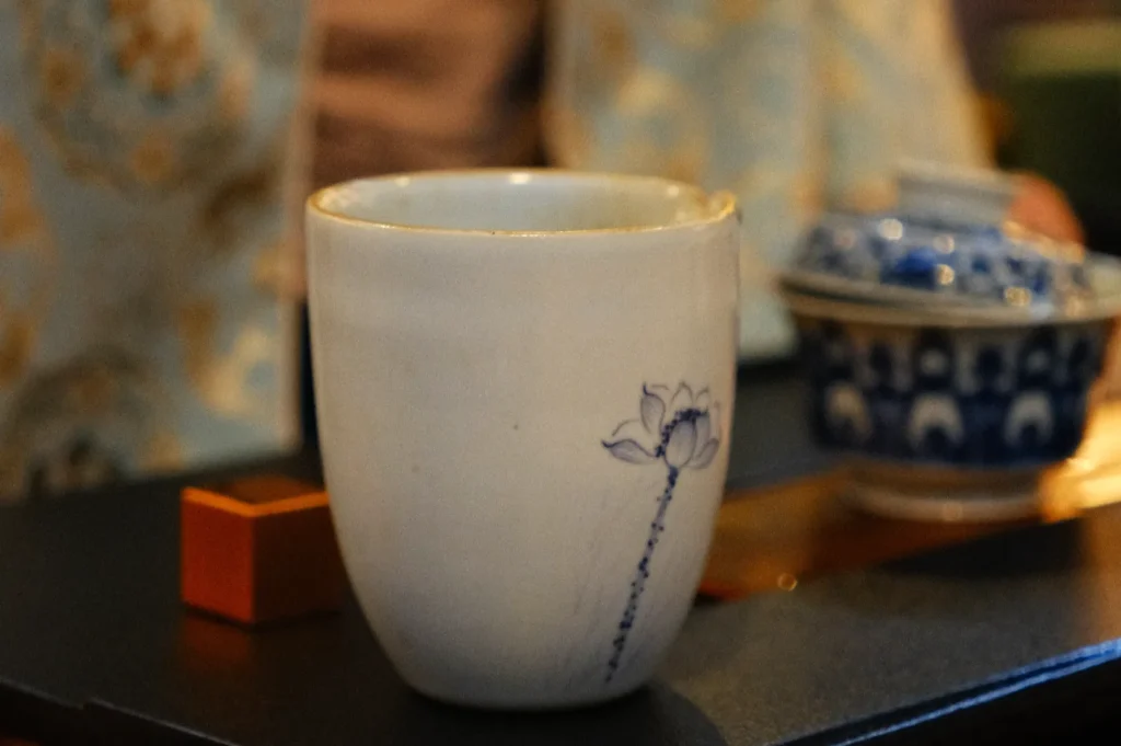 A ceramic cup with a lotus flower design, placed on a table with additional pottery in the background.