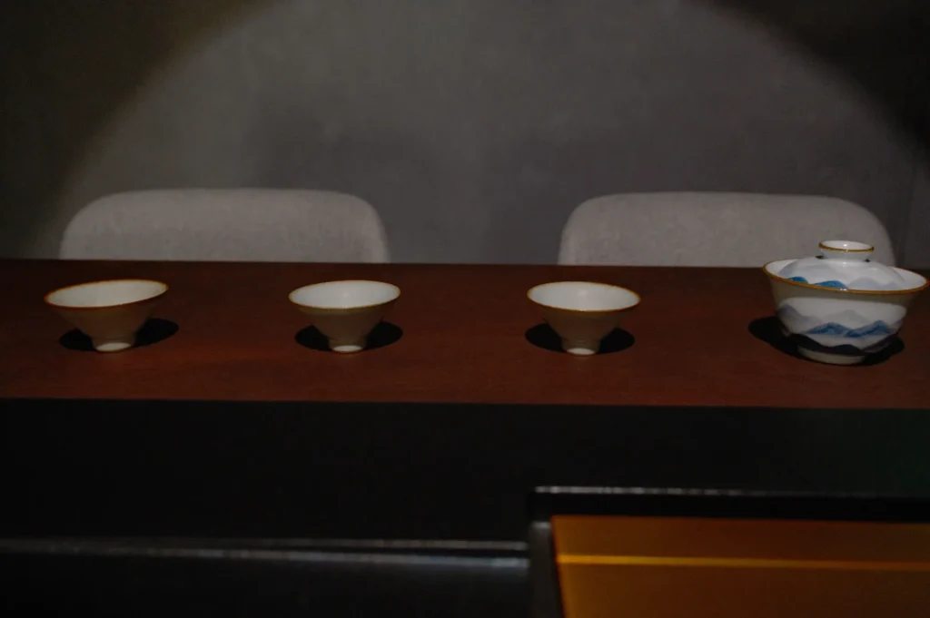 Three small, cream-colored bowls are neatly arranged on a dark wooden table, next to a lidded bowl with blue patterns. The background is softly lit.