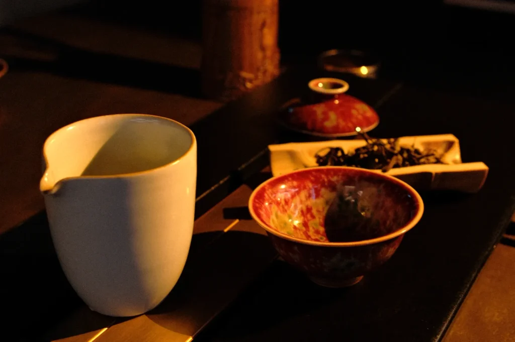 A white ceramic pitcher and a colorful red tea cup on a dark surface, with a small bamboo dish of tea leaves nearby.