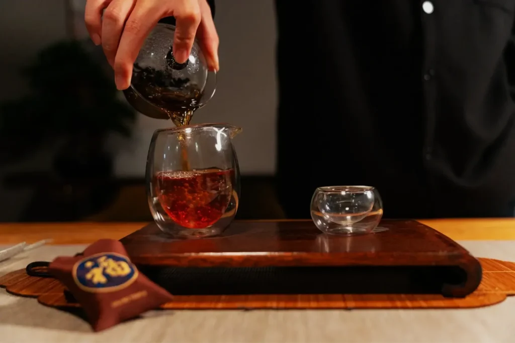 A person pouring tea from a teapot into a glass cup, with another small glass cup on a wooden tray.