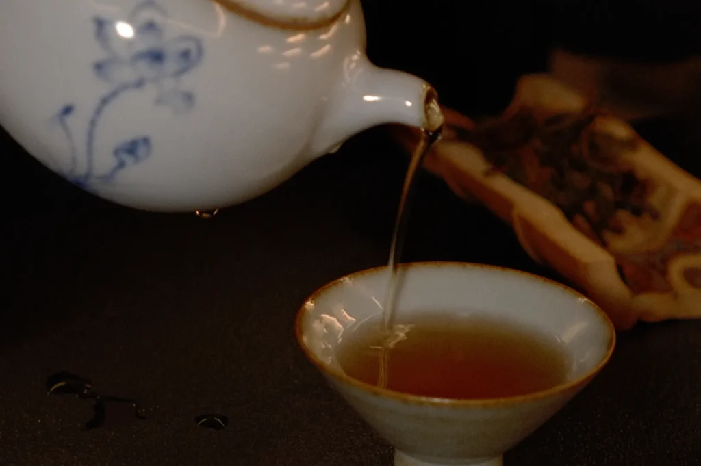 A teapot pouring tea into a small cup, with droplets falling and tea leaves placed on a half a bamboo a blurred background.