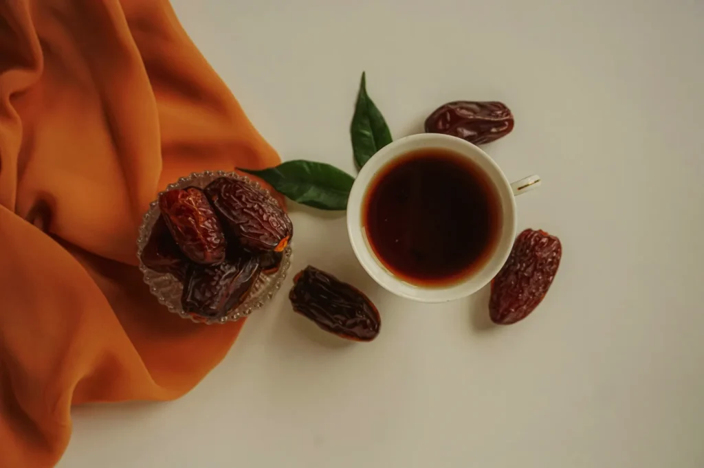 A cup of dark coffee sits next to a bowl of dates on an orange cloth. Two loose dates and a green leaf are nearby on a white background.