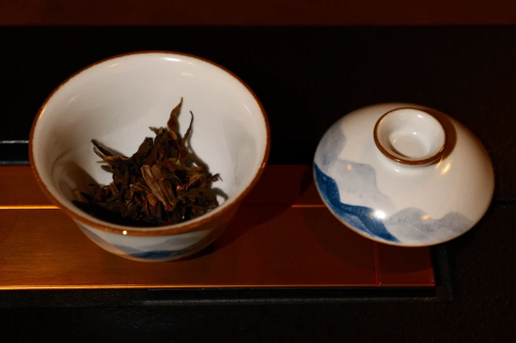 A small white bowl with dry tea leaves inside, next to a matching lid, resting on a wooden tray.