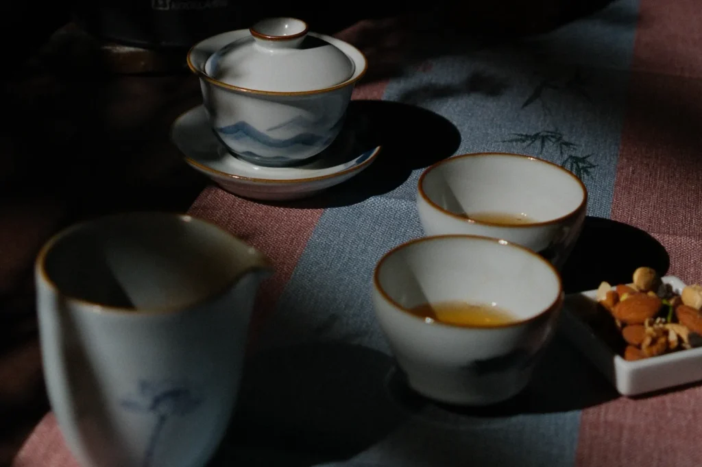 A serene tea setting with a teapot and two cups on a tablecloth, bathed in dappled sunlight. Nuts rest beside them, conveying a calm, rustic ambiance.