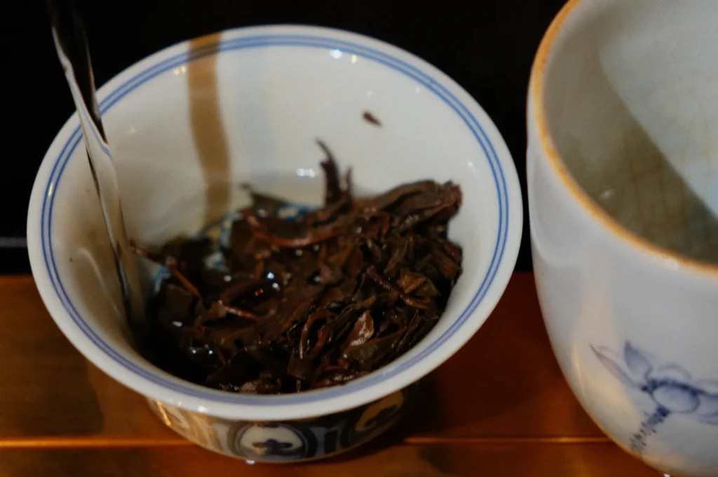A close-up of wet tea leaves in a small, decorative ceramic bowl. Hot water is being poured into the bowl, next to an empty teacup. Cozy atmosphere.