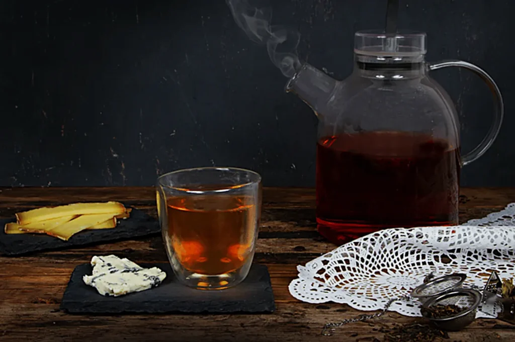A steaming glass teapot with dark tea and a double-walled glass cup sit on a rustic table. Cheese slices, blue cheese, and a lace napkin complete the cozy scene.