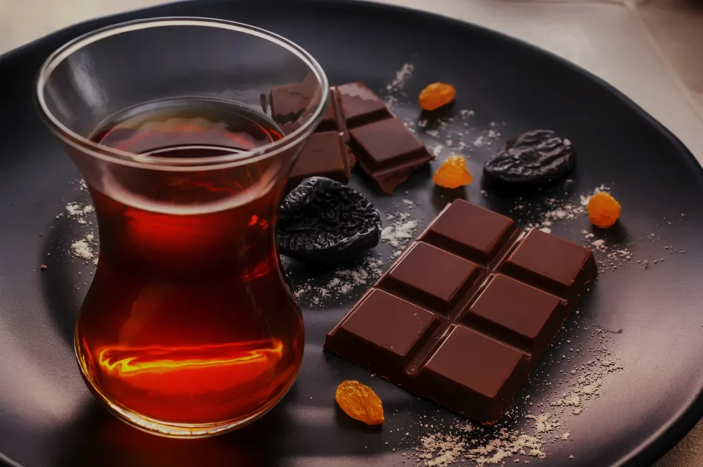 A glass of tea sits on a black plate with chocolate squares, dried prunes, and golden raisins. The setting is cozy and inviting.