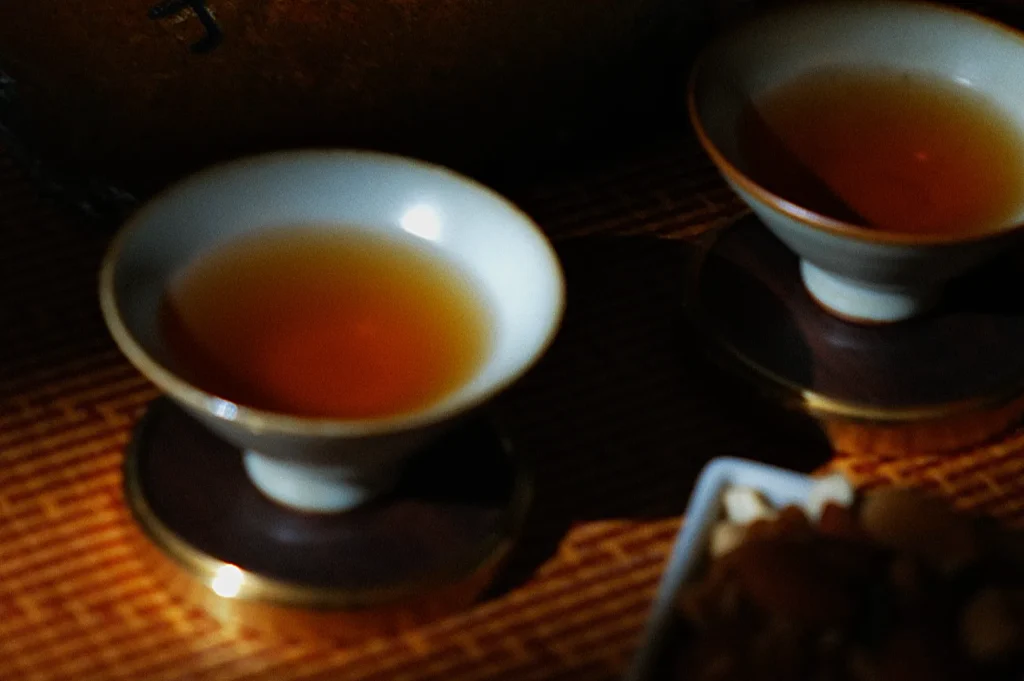 Two small porcelain cups filled with amber tea sit on dark saucers, casting gentle shadows. The warm, tranquil setting suggests a cozy tea ritual.
