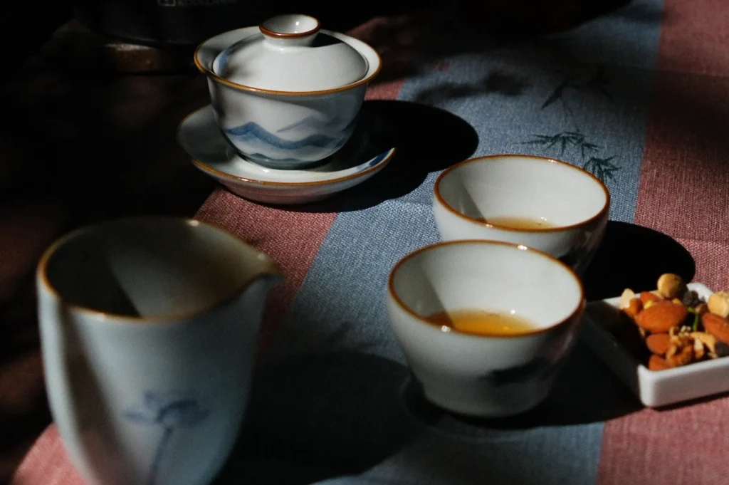 Elegant tea scene with three white ceramic teacups, a lidded teapot, and a small dish of nuts on a patterned cloth. Warm light creates a cozy, serene ambiance.