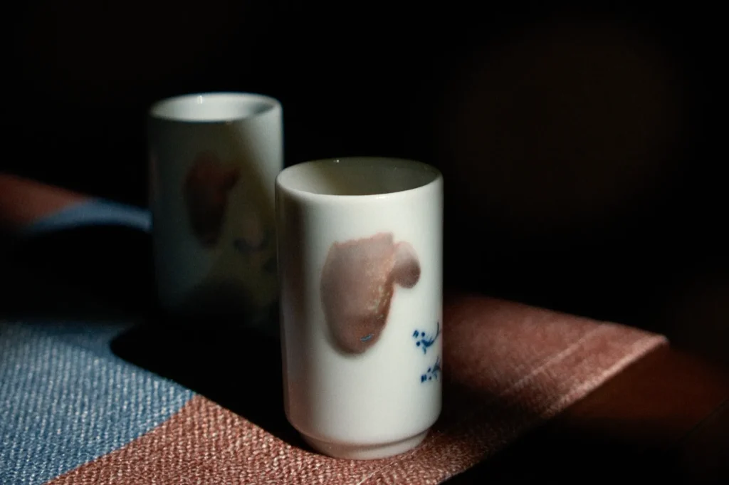 Two white ceramic cups with brown and blue designs on a textured surface, illuminated by soft light.