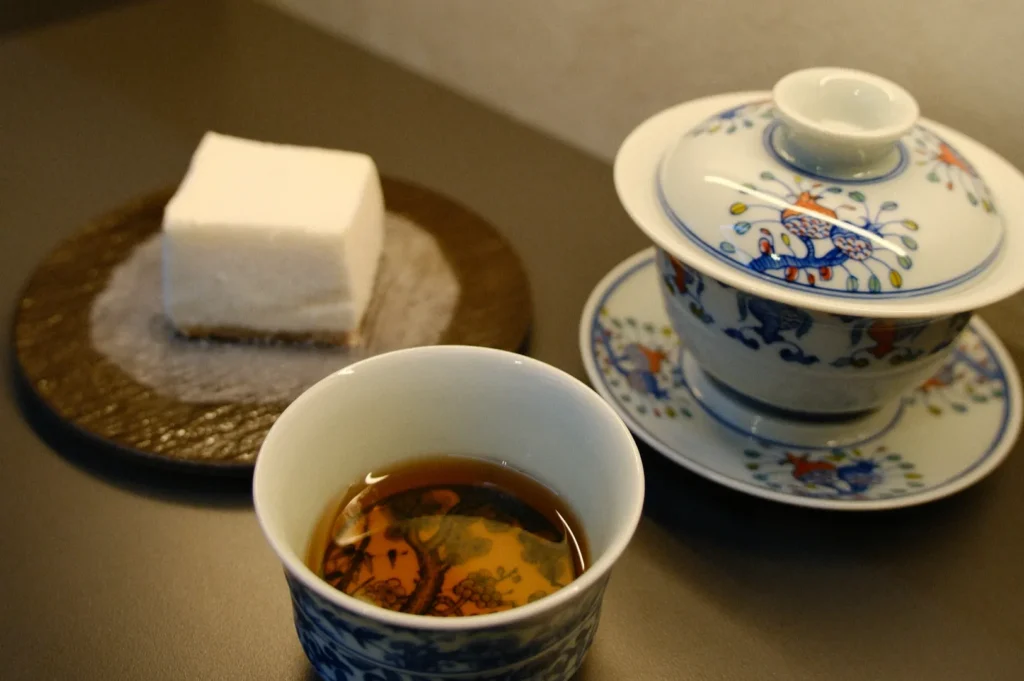 Porcelain cup with tea, patterned with colorful birds, next to a matching covered bowl. A piece of white cake on a brown plate is nearby on the table.