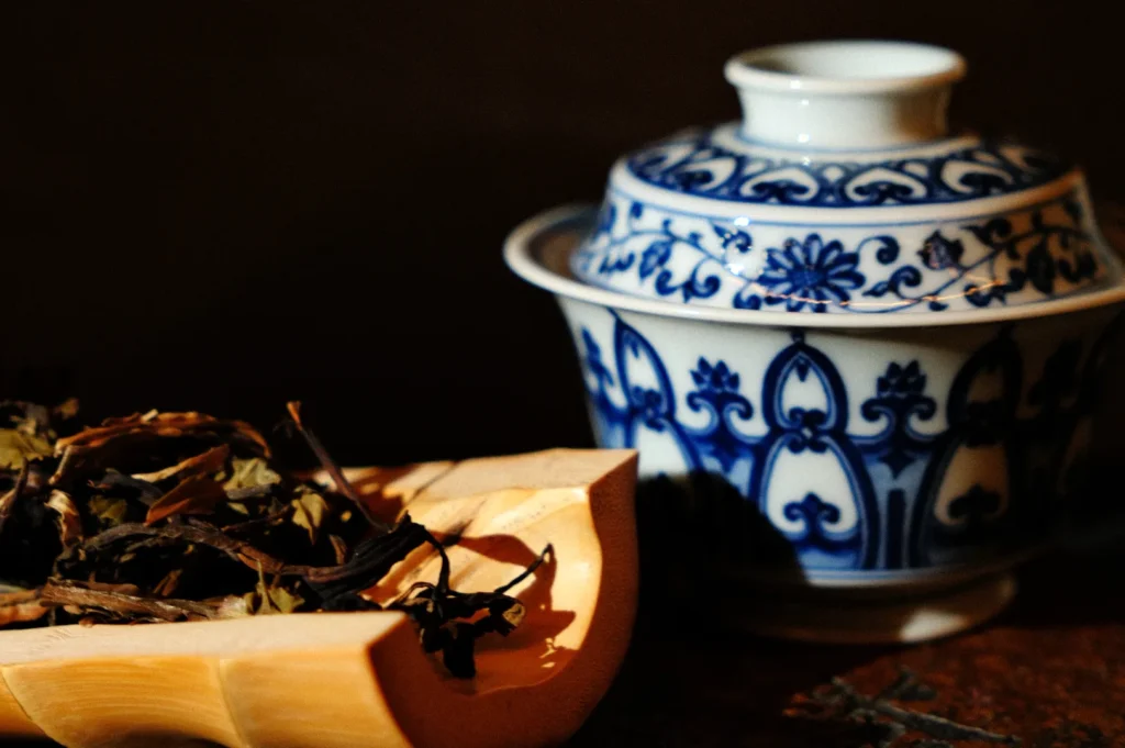 A traditional blue and white ceramic tea cup next to a bamboo container filled with dried tea leaves.
