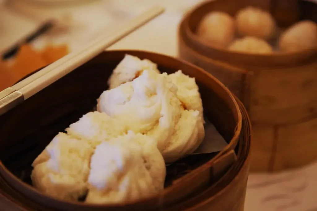 A wooden steamer basket containing fluffy white dumplings, with chopsticks resting beside it.