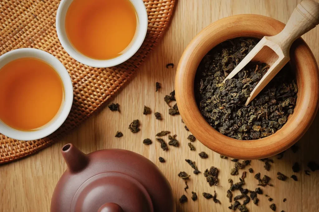 A wooden bowl filled with loose tea leaves, a wooden scoop, two cups of brewed tea, and a teapot on a wooden surface.