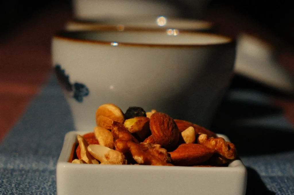 A small white dish filled with assorted nuts sits on a textured blue surface. Blurred teacups in the background create a cozy, inviting atmosphere.