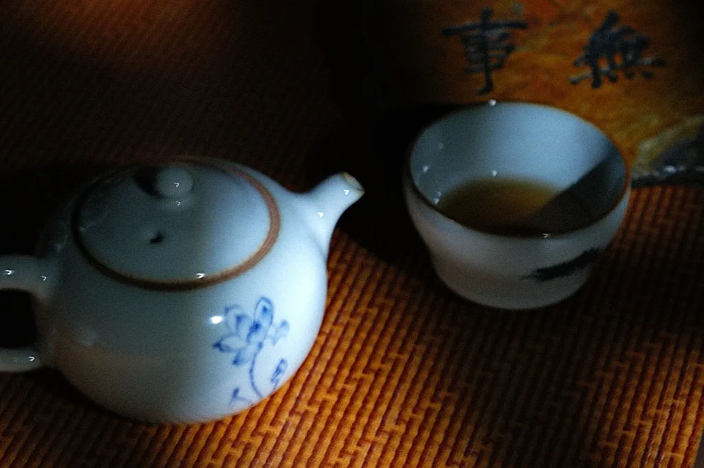 A ceramic teapot with blue floral patterns and a small cup of tea sit on a textured surface. Soft lighting creates a warm and serene atmosphere.
