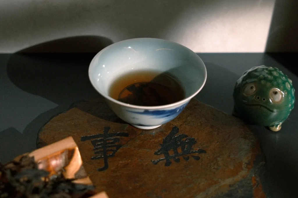 A delicate cup of amber tea sits on a dark stone with Asian characters. A green ceramic frog figurine adds a whimsical touch, evoking tranquility.