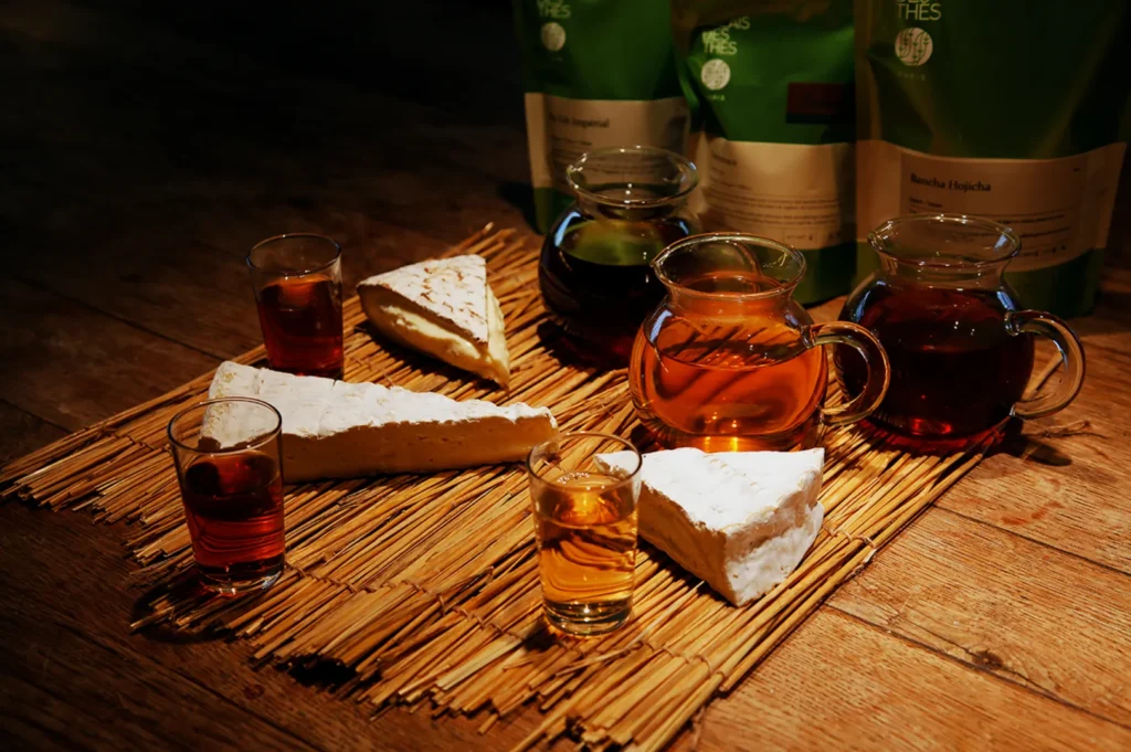 A rustic wooden table displays various teas in glass pitchers and glasses, alongside triangular pieces of brie cheese on a bamboo mat, evoking a cozy ambiance.