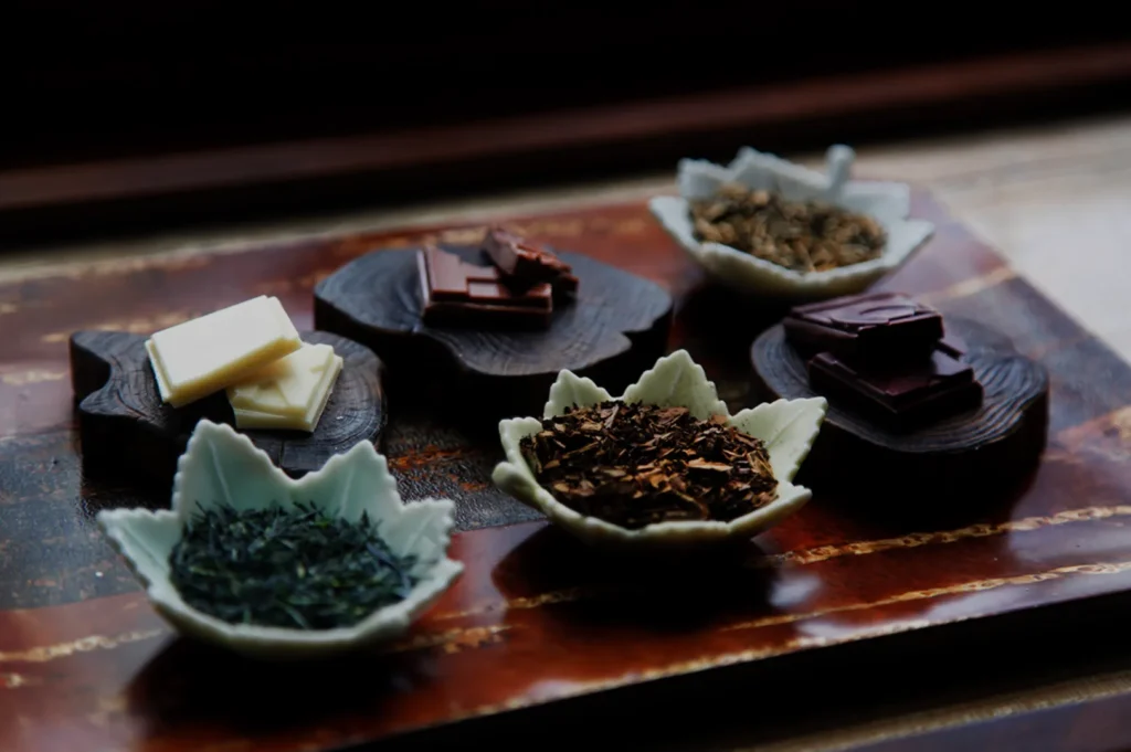 A wooden tray displays an assortment of tea leaves and chocolate, artfully arranged in small ceramic dishes. The mood is warm and inviting.