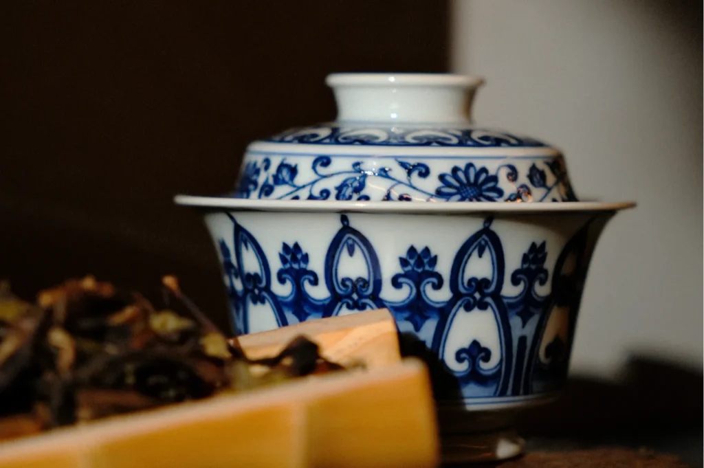 Decorative white and blue ceramic bowl with intricate patterns, partially obscured by a wooden box filled with dried leaves, evoking an elegant, serene ambiance.