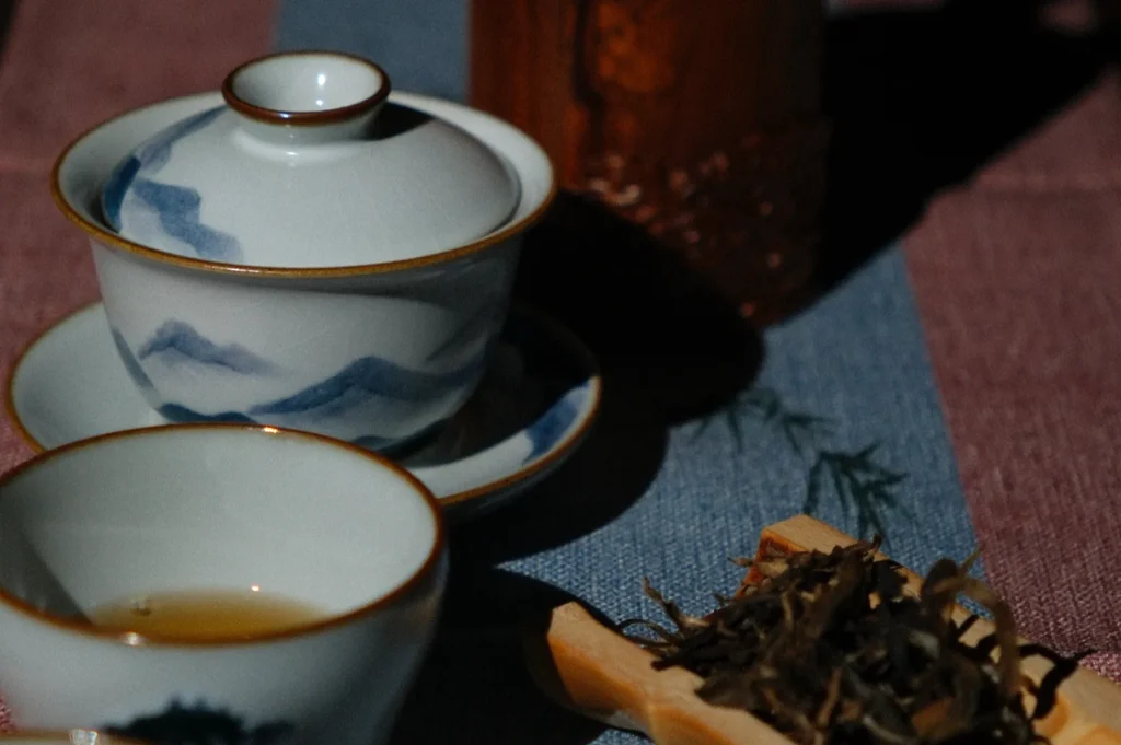 Ceramic tea set with a painted mountain design on a woven mat. Includes a teacup with tea and loose tea leaves, creating a calm, traditional ambiance.