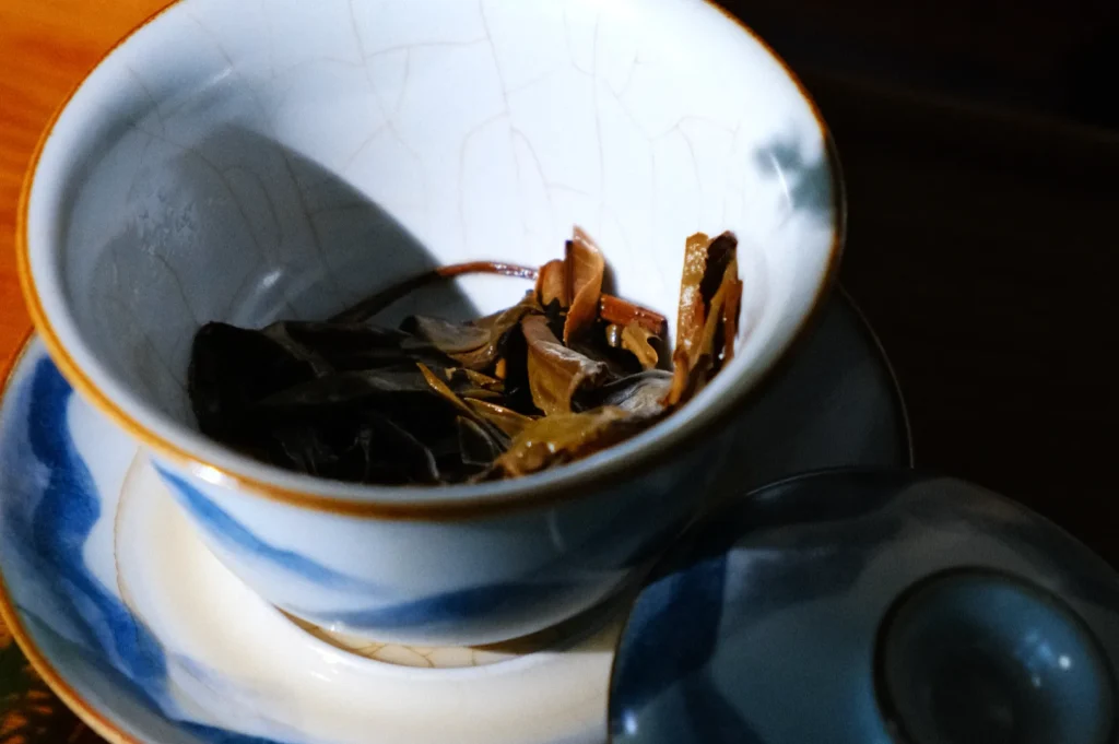 A blue and white teacup filled with dry tea leaves, placed on a matching saucer, with shadows creating contrast on the surface.