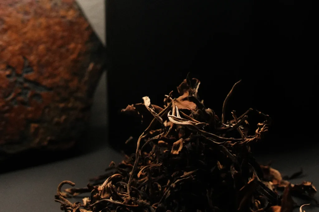 A close-up of a pile of dried leaves or herbs on a dark surface, with a wooden board and black cylindrical container in the background.