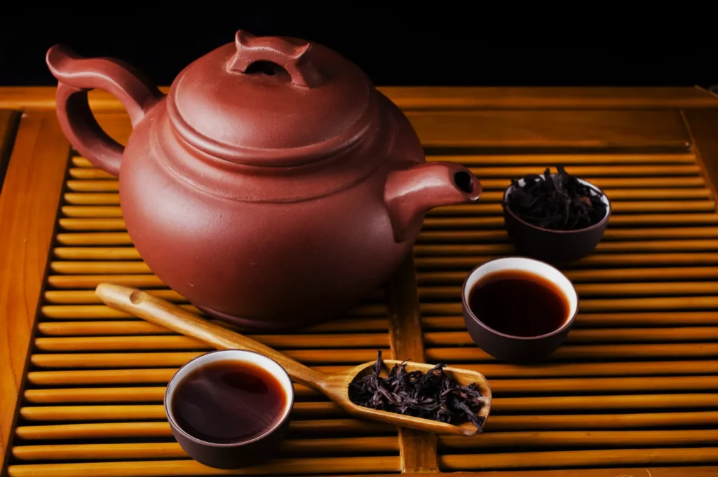 A clay teapot with two teacups filled with dark tea and bowls of loose-leaf tea resting on a bamboo mat, creating a warm, relaxing atmosphere.