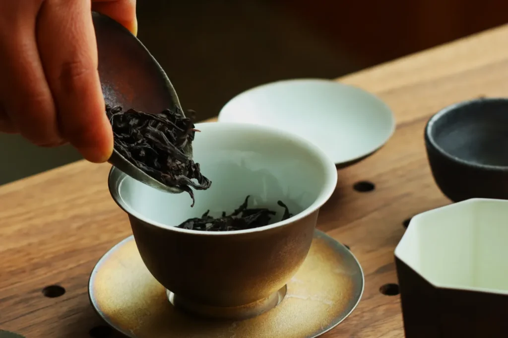 A person pouring loose tea leaves into a small cup on a wooden surface.
