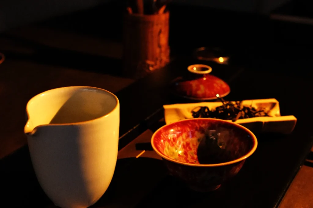 A teapot, a bowl with loose tea leaves, and a small dish are arranged on a table, illuminated by warm light.